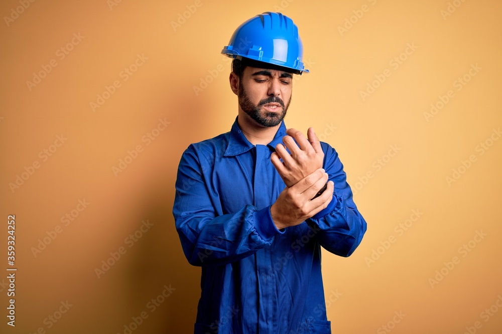 Mechanic man with beard wearing blue uniform and safety helmet over yellow background Suffering pain on hands and fingers, arthritis inflammation Mechanic man with beard wearing blue uniform and safety helmet over yellow background Suffering pain on hands and fingers, arthritis inflammation