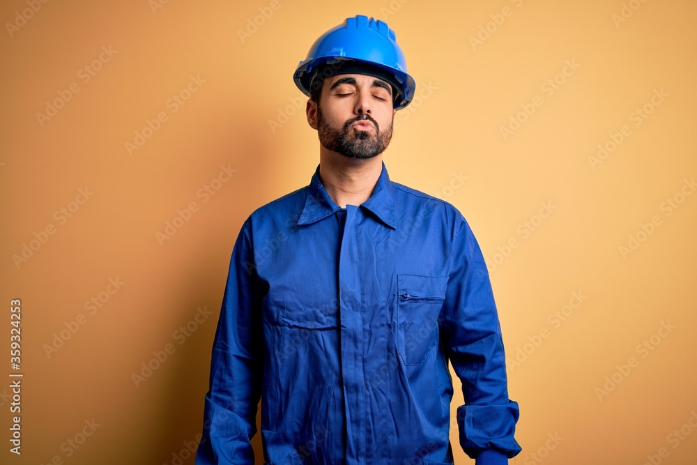 Mechanic man with beard wearing blue uniform and safety helmet over yellow background looking at the camera blowing a kiss on air being lovely and sexy. Love expression.