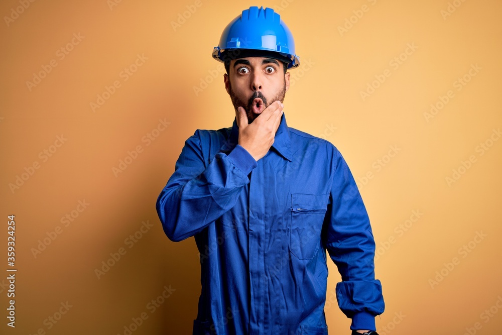 Mechanic man with beard wearing blue uniform and safety helmet over yellow background Looking fascinated with disbelief, surprise and amazed expression with hands on chin