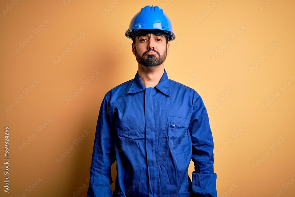 Mechanic man with beard wearing blue uniform and safety helmet over yellow background depressed and worry for distress, crying angry and afraid. Sad expression.