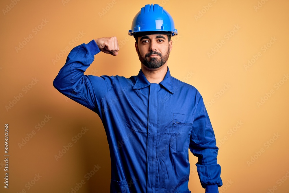 Mechanic man with beard wearing blue uniform and safety helmet over yellow background Strong person showing arm muscle, confident and proud of power