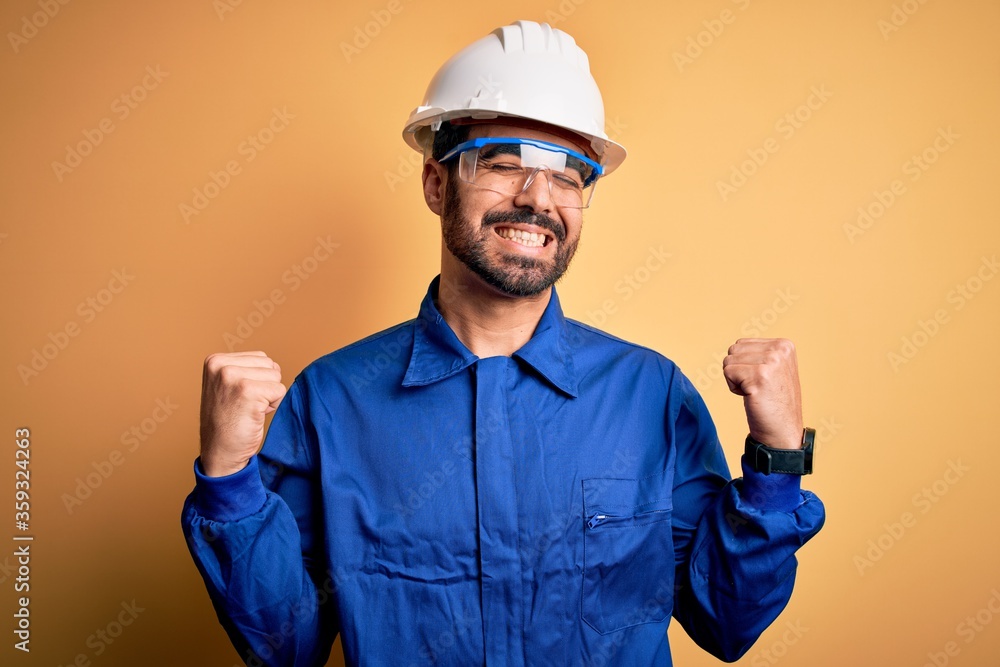 Mechanic man with beard wearing blue uniform and safety glasses over yellow background very happy and excited doing winner gesture with arms raised, smiling and screaming for success. Celebration
