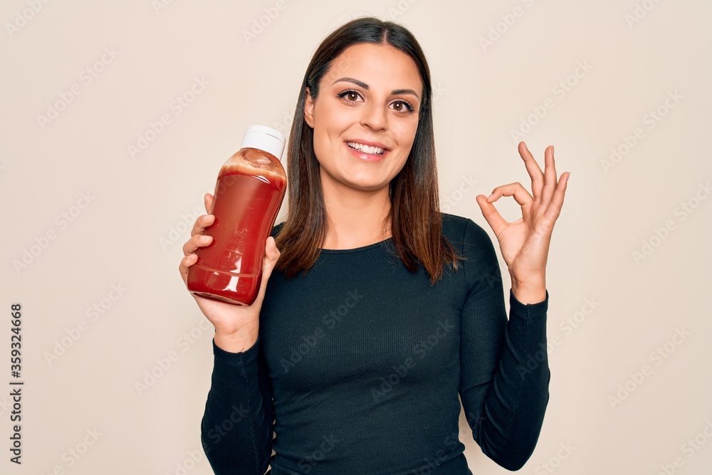 Young beautiful brunette woman holding bottle with ketchup sauce over white background doing ok sign with fingers, smiling friendly gesturing excellent symbol Young beautiful brunette woman holding bottle with ketchup sauce over white background doing ok sign with fingers, smiling friendly gesturing excellent symbol