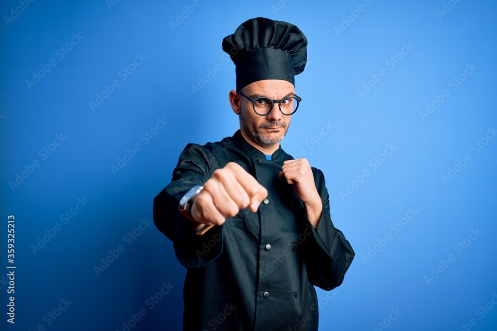Young handsome chef man wearing cooker uniform and hat over isolated blue background Punching fist to fight, aggressive and angry attack, threat and violence Young handsome chef man wearing cooker uniform and hat over isolated blue background Punching fist to fight, aggressive and angry attack, threat and violence