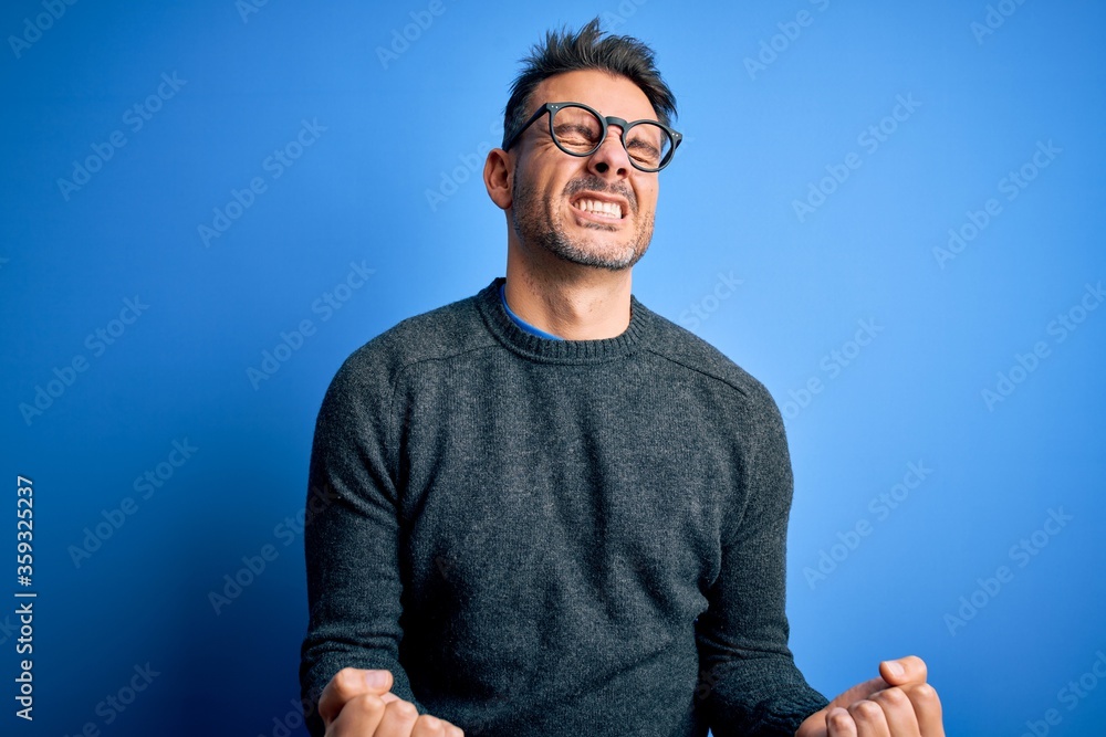 Young handsome man wearing casual sweater and glasses standing over blue background very happy and excited doing winner gesture with arms raised, smiling and screaming for success. Celebration Young handsome man wearing casual sweater and glasses standing over blue background very happy and excited doing winner gesture with arms raised, smiling and screaming for success. Celebration