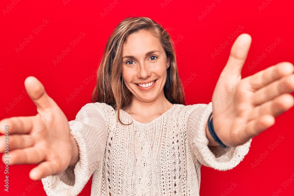 Young beautiful blonde woman wearing winter sweater over red isolated background looking at the camera smiling with open arms for hug. cheerful expression embracing happiness. Young beautiful blonde woman wearing winter sweater over red isolated background looking at the camera smiling with open arms for hug. cheerful expression embracing happiness.