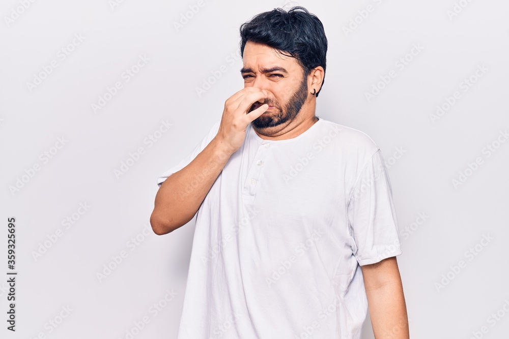 Young hispanic man wearing casual clothes smelling something stinky and disgusting, intolerable smell, holding breath with fingers on nose. bad smell