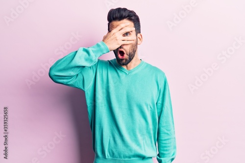 Young handsome man with beard wearing casual sweater peeking in shock covering face and eyes with hand, looking through fingers afraid © Krakenimages.com