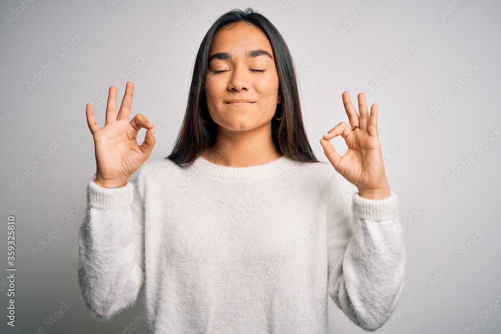 Young beautiful asian woman wearing casual sweater standing over white background relax and smiling with eyes closed doing meditation gesture with fingers. Yoga concept. Young beautiful asian woman wearing casual sweater standing over white background relax and smiling with eyes closed doing meditation gesture with fingers. Yoga concept.
