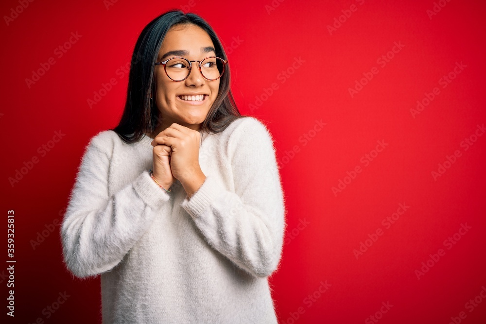 Young beautiful asian woman wearing casual sweater and glasses over red background laughing nervous and excited with hands on chin looking to the side Young beautiful asian woman wearing casual sweater and glasses over red background laughing nervous and excited with hands on chin looking to the side