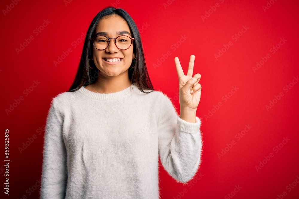 Young beautiful asian woman wearing casual sweater and glasses over red background showing and pointing up with fingers number two while smiling confident and happy. Young beautiful asian woman wearing casual sweater and glasses over red background showing and pointing up with fingers number two while smiling confident and happy.