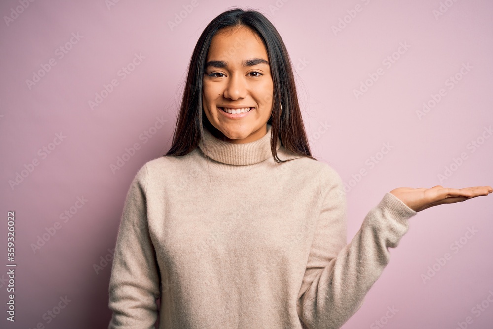Young beautiful asian woman wearing casual turtleneck sweater over pink background smiling cheerful presenting and pointing with palm of hand looking at the camera. Young beautiful asian woman wearing casual turtleneck sweater over pink background smiling cheerful presenting and pointing with palm of hand looking at the camera.