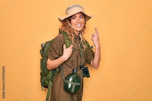 Photo Young blonde explorer woman with blue eyes hiking wearing backpack and water canteen smiling amazed and surprised and pointing up with fingers and raised arms
