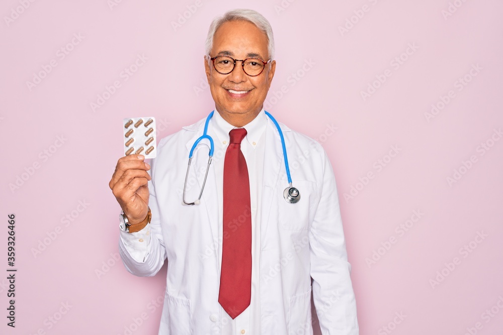 Middle age senior grey-haired doctor man holding prescription pills over pink background with a happy face standing and smiling with a confident smile showing teeth Middle age senior grey-haired doctor man holding prescription pills over pink background with a happy face standing and smiling with a confident smile showing teeth