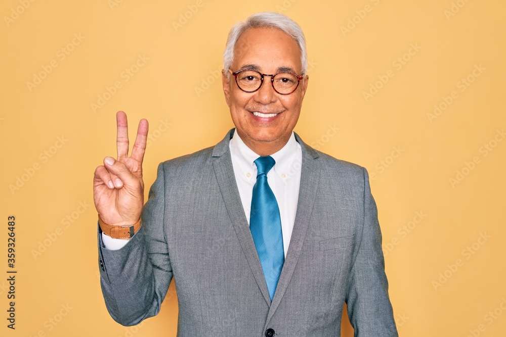 Middle age senior grey-haired handsome business man wearing glasses over yellow background showing and pointing up with fingers number two while smiling confident and happy. Middle age senior grey-haired handsome business man wearing glasses over yellow background showing and pointing up with fingers number two while smiling confident and happy.