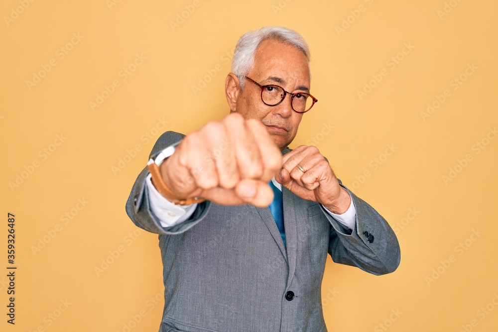 Middle age senior grey-haired handsome business man wearing glasses over yellow background Punching fist to fight, aggressive and angry attack, threat and violence Middle age senior grey-haired handsome business man wearing glasses over yellow background Punching fist to fight, aggressive and angry attack, threat and violence