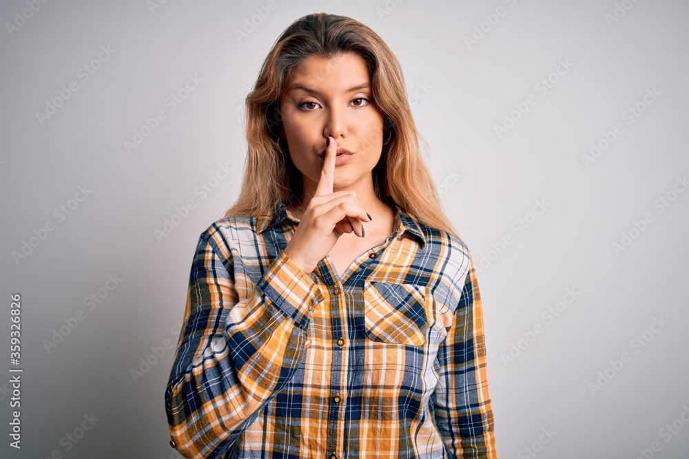 Young beautiful blonde woman wearing casual shirt standing over isolated white background asking to be quiet with finger on lips. Silence and secret concept.
