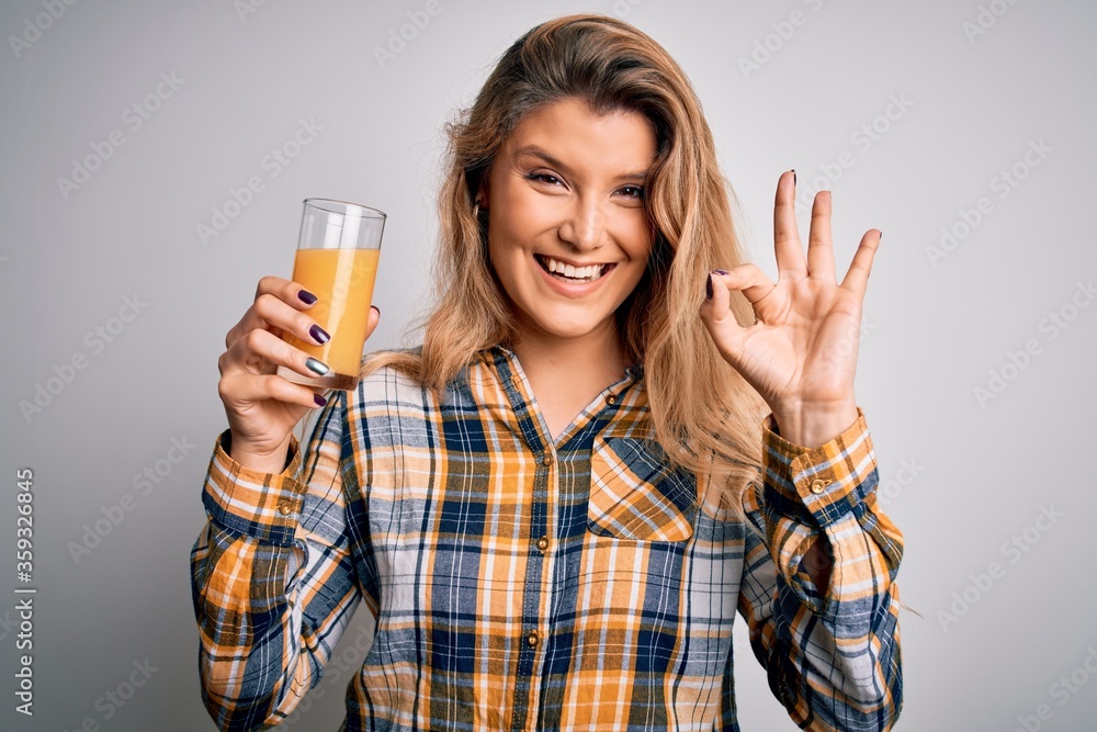 Young beautiful blonde woman drinking glass of healthy orange juice over white background doing ok sign with fingers, excellent symbol Young beautiful blonde woman drinking glass of healthy orange juice over white background doing ok sign with fingers, excellent symbol