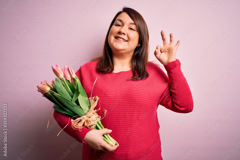 Beautiful plus size woman holding romantic bouquet of natural tulips flowers over pink background smiling positive doing ok sign with hand and fingers. Successful expression. Beautiful plus size woman holding romantic bouquet of natural tulips flowers over pink background smiling positive doing ok sign with hand and fingers. Successful expression.