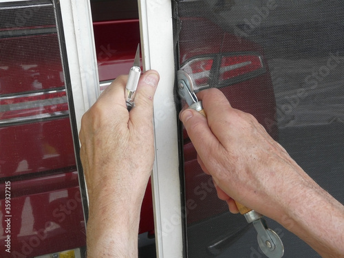Repairing Screen Door with Spline Tool. A homeowner is using a spline roller tool and knife to replace a damaged panel in a sliding garage screen door with a metal frame.