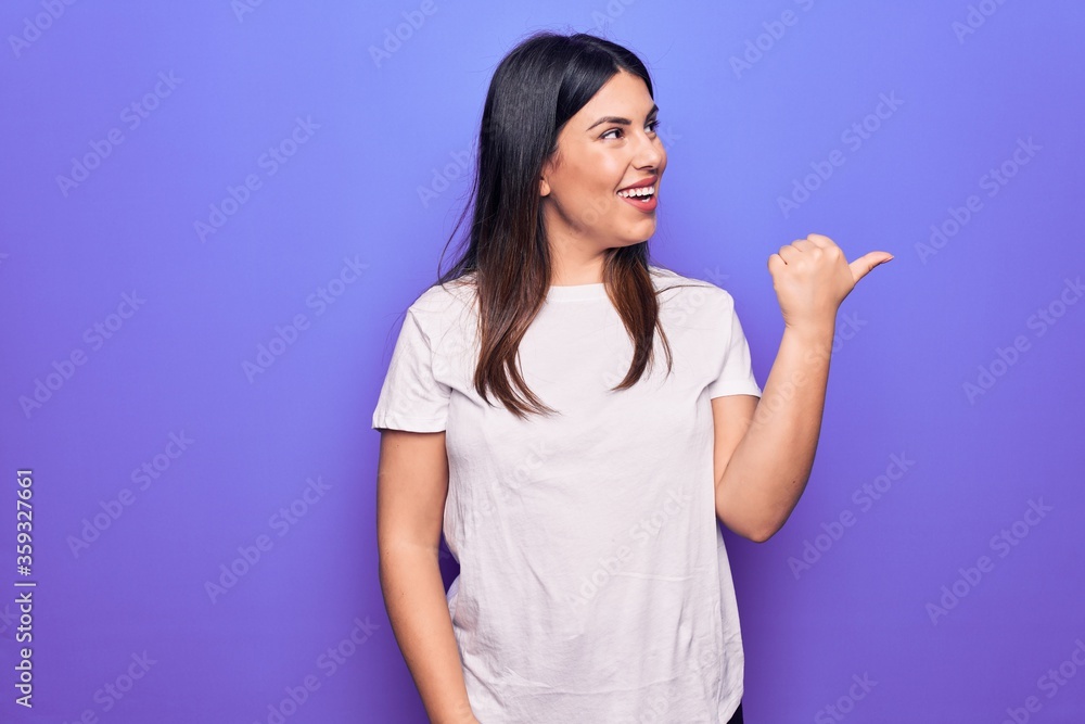Young beautiful brunette woman wearing casual t-shirt standing over purple background pointing thumb up to the side smiling happy with open mouth Young beautiful brunette woman wearing casual t-shirt standing over purple background pointing thumb up to the side smiling happy with open mouth
