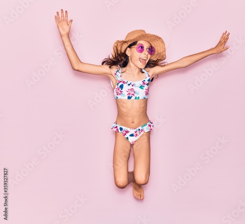 Adorable hispanic child girl on vacation wearing bikini, sunglasses and hat smiling happy. Jumping with smile on face over isolated pink background