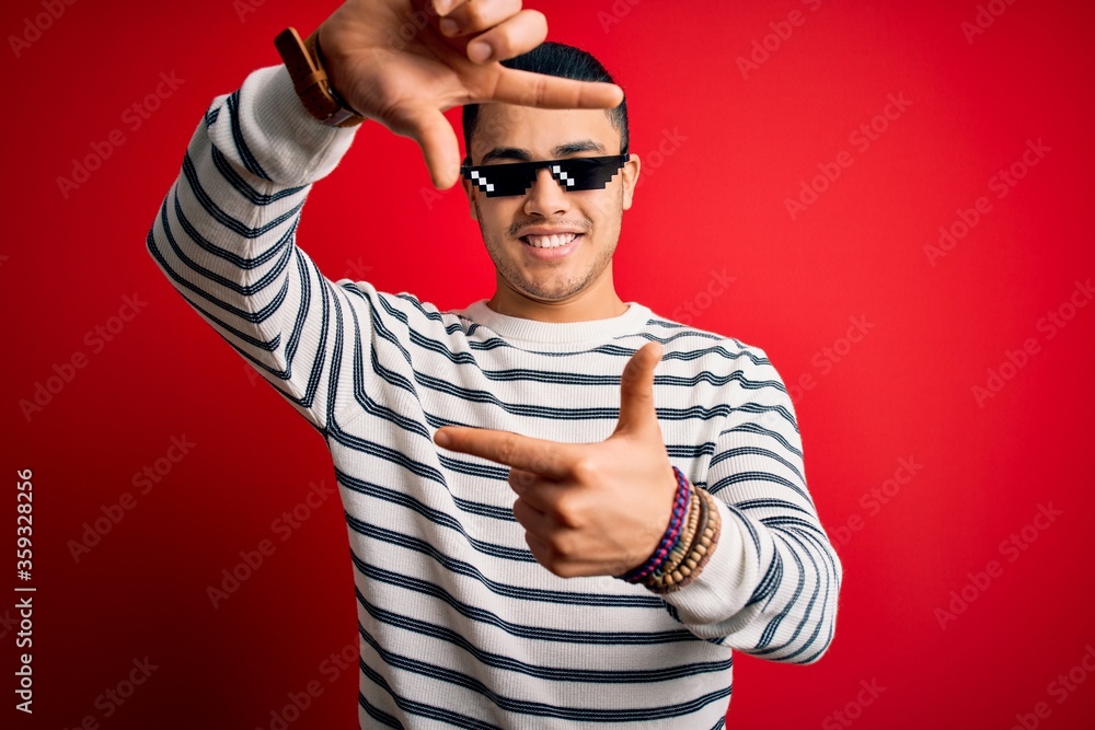 Naklejka premium Young brazilian man wearing funny thug life sunglasses over isolated red background smiling making frame with hands and fingers with happy face. Creativity and photography concept.