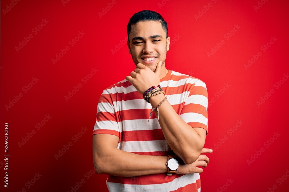 Young brazilian man wearing casual striped t-shirt standing over isolated red background looking confident at the camera smiling with crossed arms and hand raised on chin. Thinking positive. Young brazilian man wearing casual striped t-shirt standing over isolated red background looking confident at the camera smiling with crossed arms and hand raised on chin. Thinking positive.
