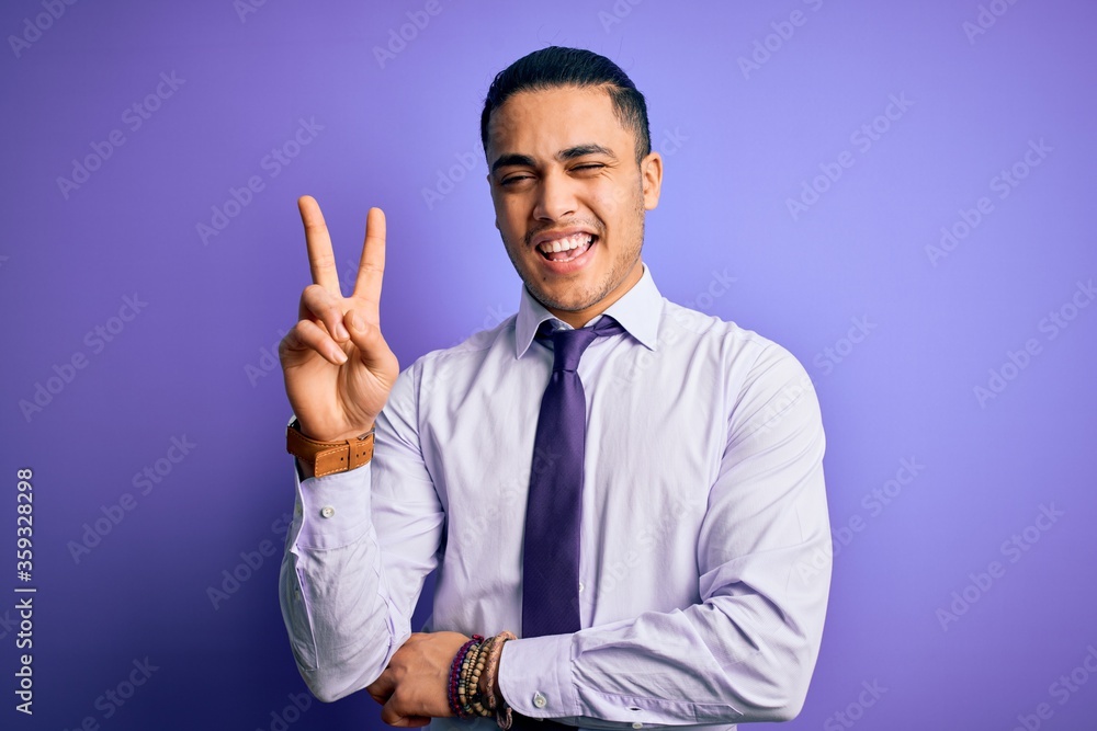 Young brazilian businessman wearing elegant tie standing over isolated purple background smiling with happy face winking at the camera doing victory sign with fingers. Number two. Young brazilian businessman wearing elegant tie standing over isolated purple background smiling with happy face winking at the camera doing victory sign with fingers. Number two.