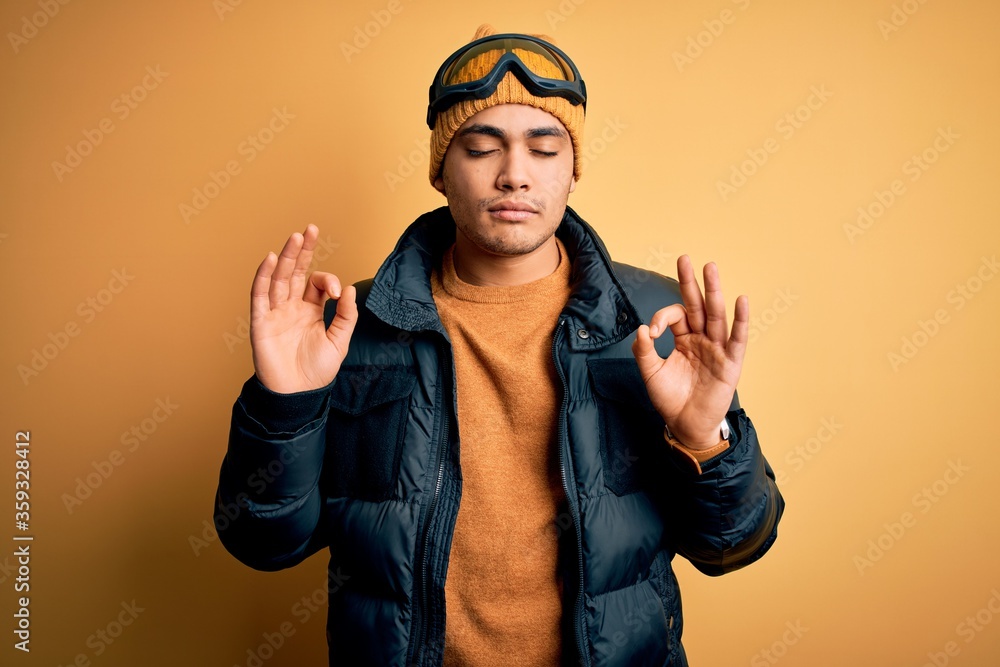 Young brazilian skier man wearing snow sportswear and ski goggles over yellow background relaxed and smiling with eyes closed doing meditation gesture with fingers. Yoga concept. Young brazilian skier man wearing snow sportswear and ski goggles over yellow background relaxed and smiling with eyes closed doing meditation gesture with fingers. Yoga concept.