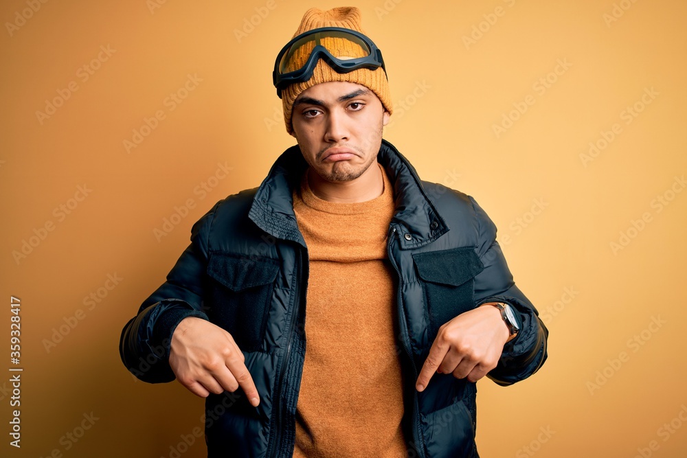 Young brazilian skier man wearing snow sportswear and ski goggles over yellow background Pointing down looking sad and upset, indicating direction with fingers, unhappy and depressed. Young brazilian skier man wearing snow sportswear and ski goggles over yellow background Pointing down looking sad and upset, indicating direction with fingers, unhappy and depressed.