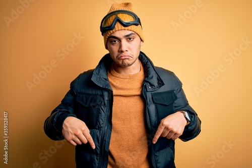 Young brazilian skier man wearing snow sportswear and ski goggles over yellow background Pointing down looking sad and upset, indicating direction with fingers, unhappy and depressed. © Krakenimages.com