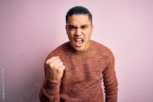 Young brazilian man wearing casual sweater standing over isolated pink background angry and mad raising fist frustrated and furious while shouting with anger. Rage and aggressive concept.