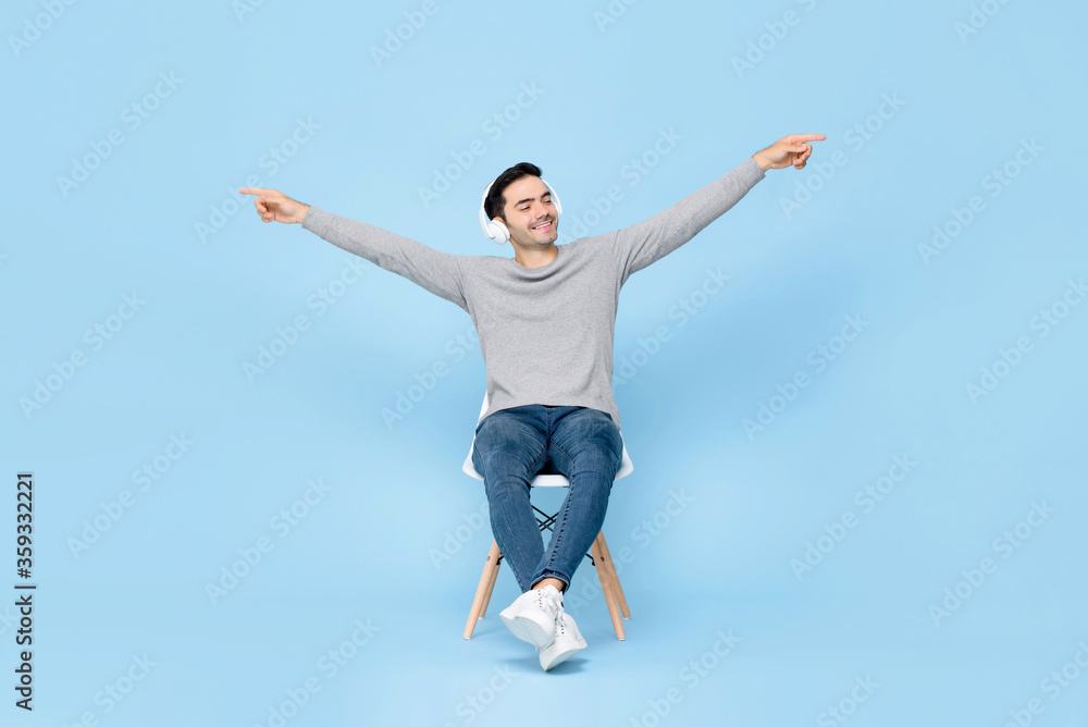 Portrait of relaxing young handsome Caucasian man sitting on chair wearing headphone listening to music with arms raised in isolated studio blue background Portrait of relaxing young handsome Caucasian man sitting on chair wearing headphone listening to music with arms raised in isolated studio blue background