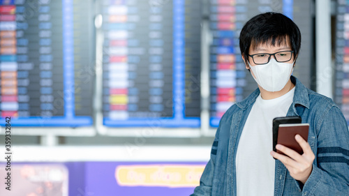 Asian man tourist wearing face mask holding smartphone and passport at arrival departure board in airport terminal. Coronavirus (COVID-19) pandemic prevention when travel. Social distancing concept