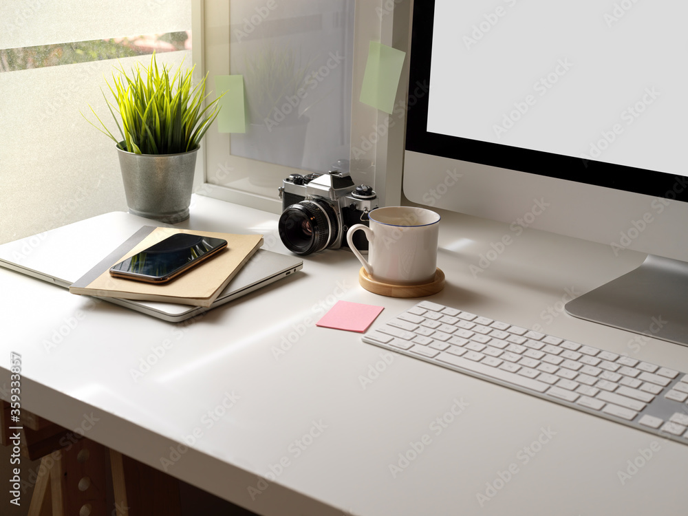 Fototapeta premium Modern office desk with mock-up computer, smartphone, camera and office supplies beside window
