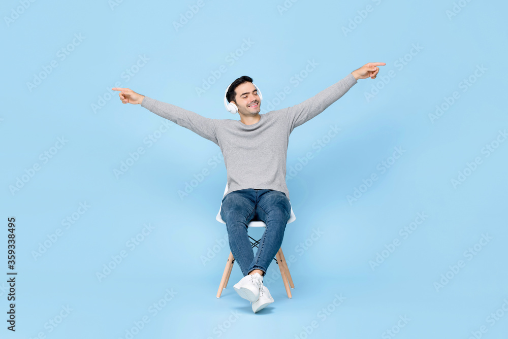 Portrait of relaxing young handsome Caucasian man sitting on chair wearing headphone listening to music with arms raised in isolated studio blue background