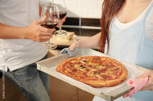 Cropped image of young woman bringing tray with freshly backed pizza to table when her boyfriend carrying glasses of red wine