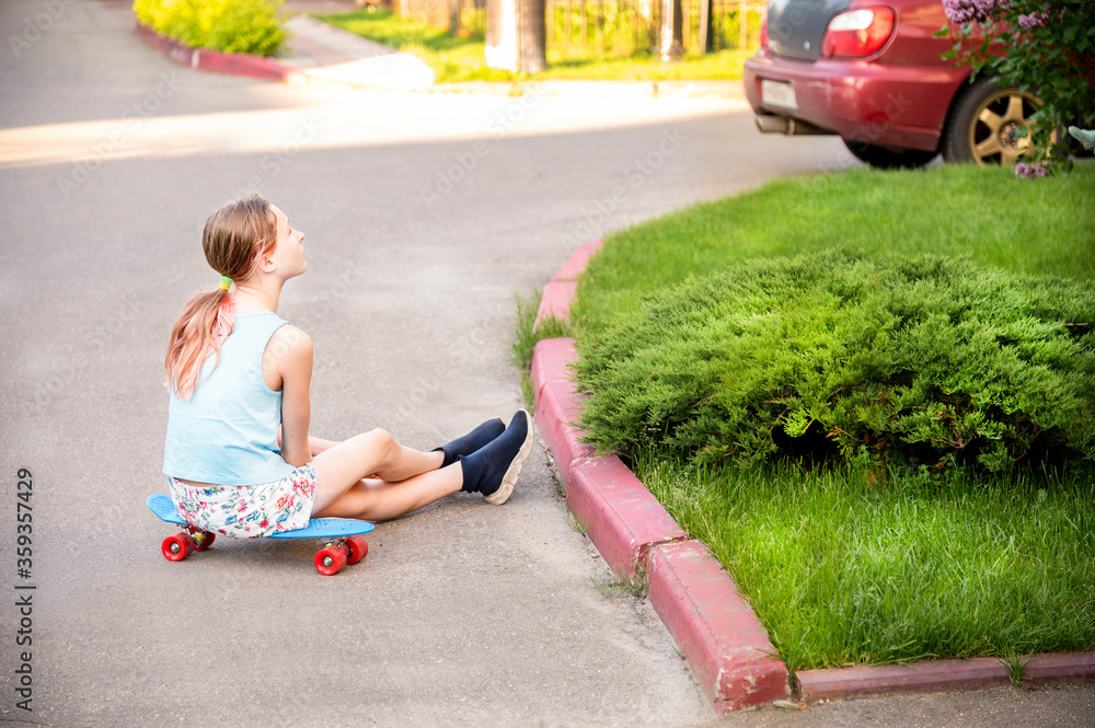 Teen girl is on the street. She is dressed in a blue T-shirt, shorts in a multi-colored flower, blue boots on her feet. The girl is sitting on a wakeboard, resting and looking thoughtfully Teen girl is on the street. She is dressed in a blue T-shirt, shorts in a multi-colored flower, blue boots on her feet. The girl is sitting on a wakeboard, resting and looking thoughtfully