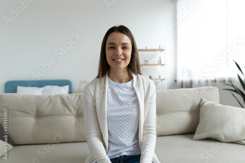 Pretty woman sitting on sofa in living room smile look at camera taking part videocall involved in virtual conferencing with colleagues, passing job interview distantly from home. Video event concept