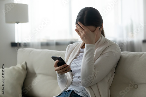 Woman sit on sofa holding smartphone cover face with hand feels scared humiliated suffering from cyberbullying being on-line abused by stalker. Bad news, life troubles, break up with boyfriend concept photo
