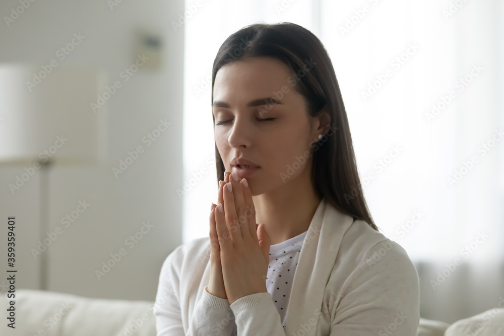 Sincere candid young woman pious deeply religious girl joined hands praying say prayer sit on sofa in living room at home. Faith in God and religion, pleading for the innermost, morning ritual concept Sincere candid young woman pious deeply religious girl joined hands praying say prayer sit on sofa in living room at home. Faith in God and religion, pleading for the innermost, morning ritual concept