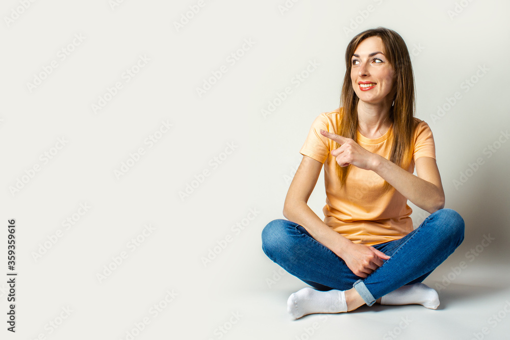 Young woman in casual clothes looks away and points a finger while sitting on the floor on a light background Young woman in casual clothes looks away and points a finger while sitting on the floor on a light background