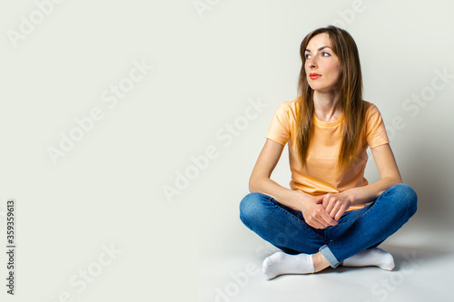 Young woman in casual clothes looks away sitting on the floor on a light background