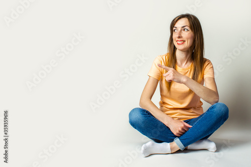 Young woman in casual clothes looks away and points a finger while sitting on the floor on a light background