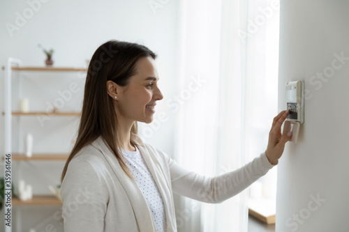 Tableau sur toile Smiling woman standing near wall-mounted device adjusting degrees in living room set comfortable temperature using thermostat home heating system