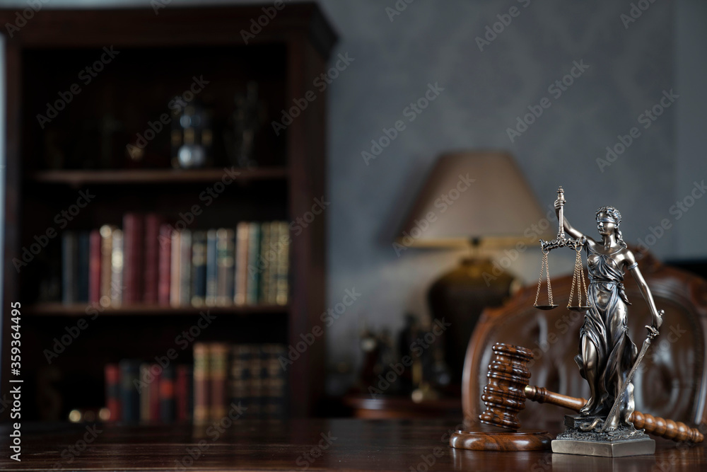 Judge chamber. Gavel and Themis statue on brown shining desk. Collection of legal books in the bookshelf.