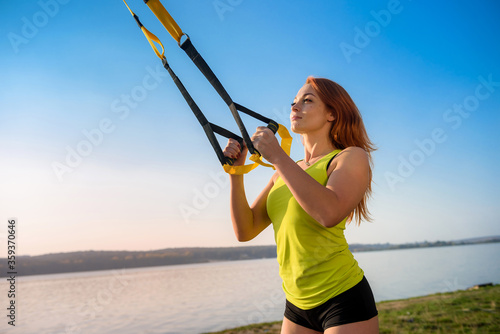 Attractive young woman doing TRX training outdoors near the lake at daytime. Healthy lifestyle.