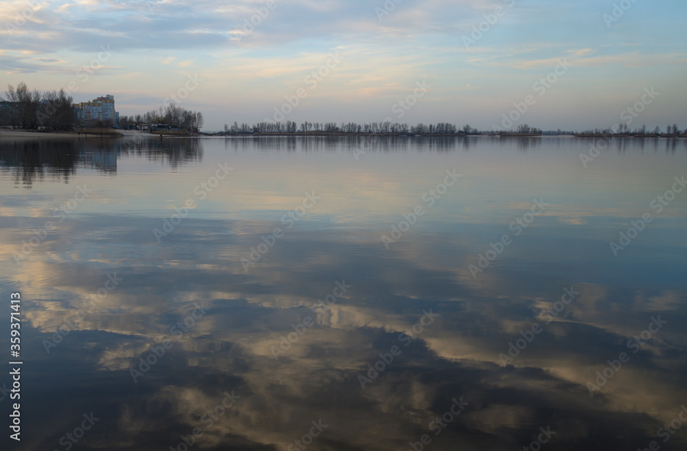 Landscape in the bay at the Dnieper river, Ukraine. Reflection of blue sky and light pink clouds in calm water. Buildings and trees on the shore on the horizon. Quiet March spring evening.