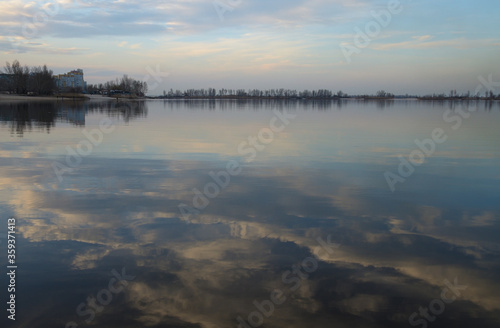 Landscape in the bay at the Dnieper river, Ukraine. Reflection of blue sky and light pink clouds in calm water. Buildings and trees on the shore on the horizon. Quiet March spring evening.